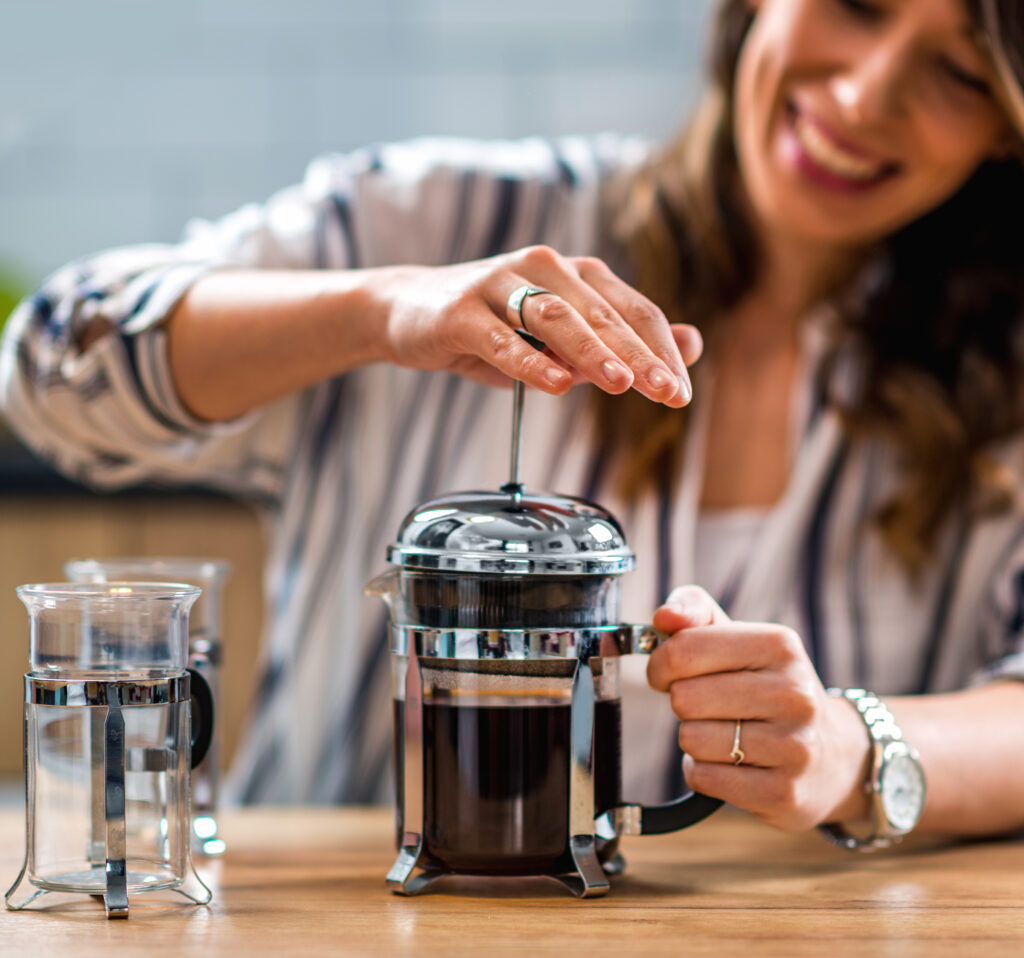 A smiling woman perfecting her French press brewing technique, pressing the plunger on a stainless steel French press for an even flavor extraction, representing the personal touch in crafting a nuanced coffee flavor with an eco-friendly method.
